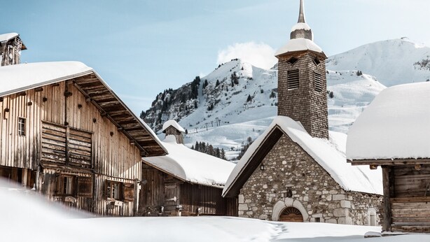 A rustic wooden building with a stone church tower, surrounded by snow-covered mountains.
