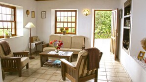 Un salon chaleureux avec un canapé, des fauteuils, une table basse et une bibliothèque. Il y a des fenêtres avec des rideaux, une lampe et une porte qui donne sur l’extérieur.