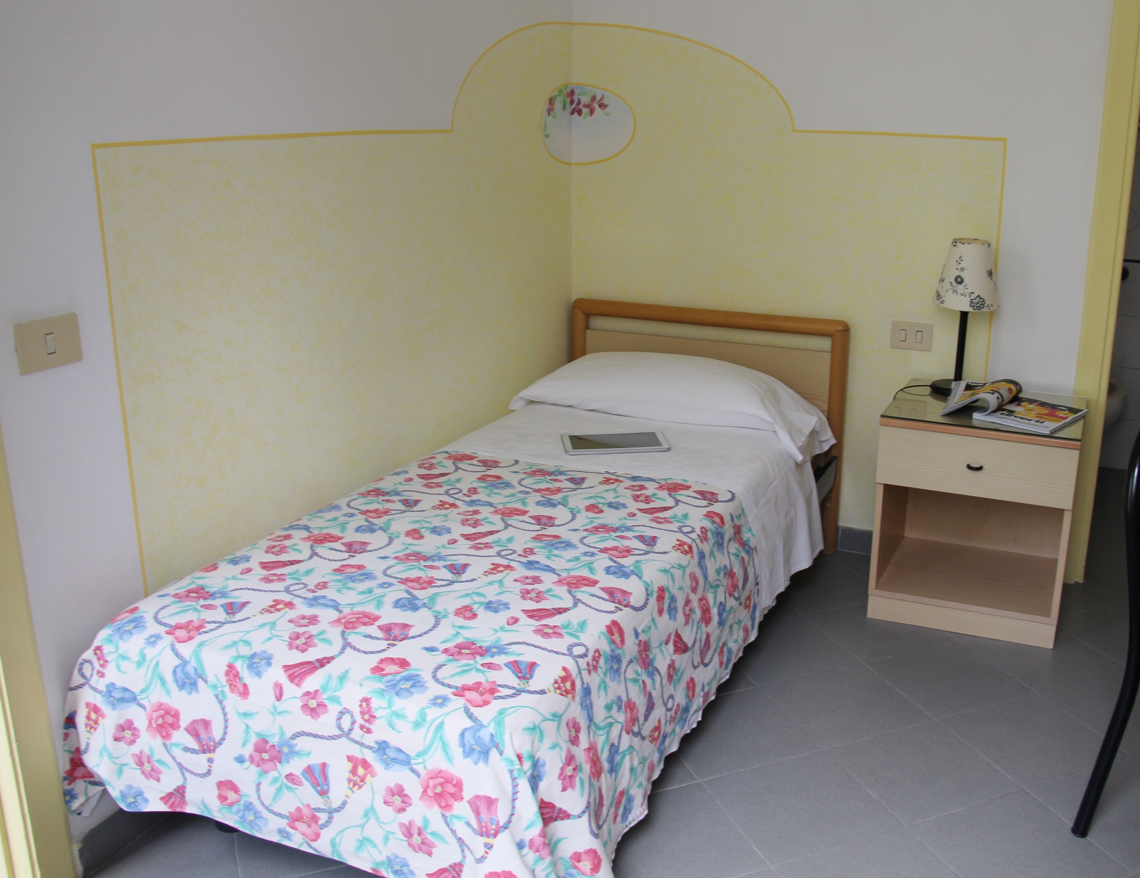 Un petit lit bien fait, avec une couvre-lit à motifs floraux, une table de chevet avec une lampe et une petite commode.