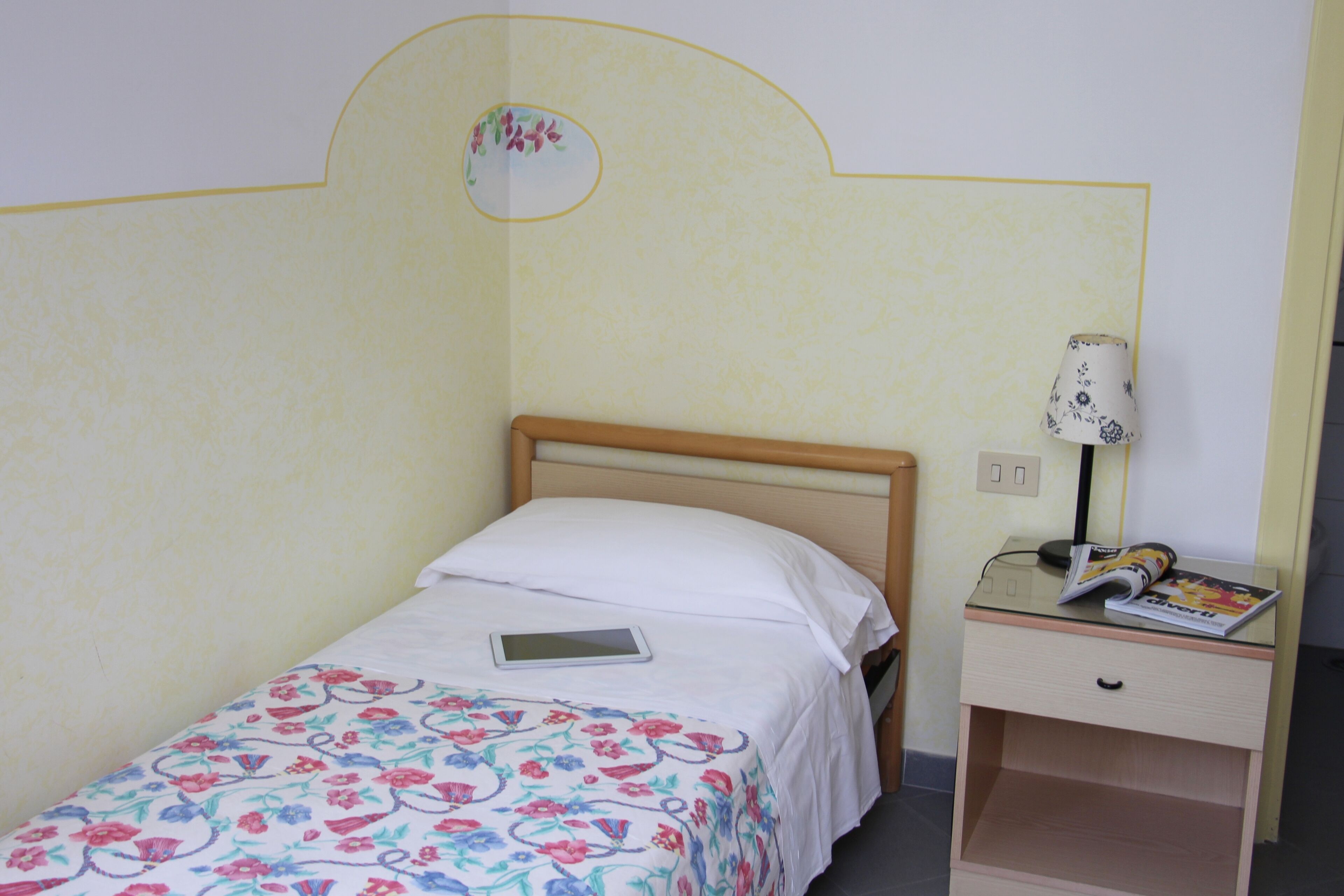 Una cama individual con colcha estampada, una mesita de noche con lámpara y una revista encima.