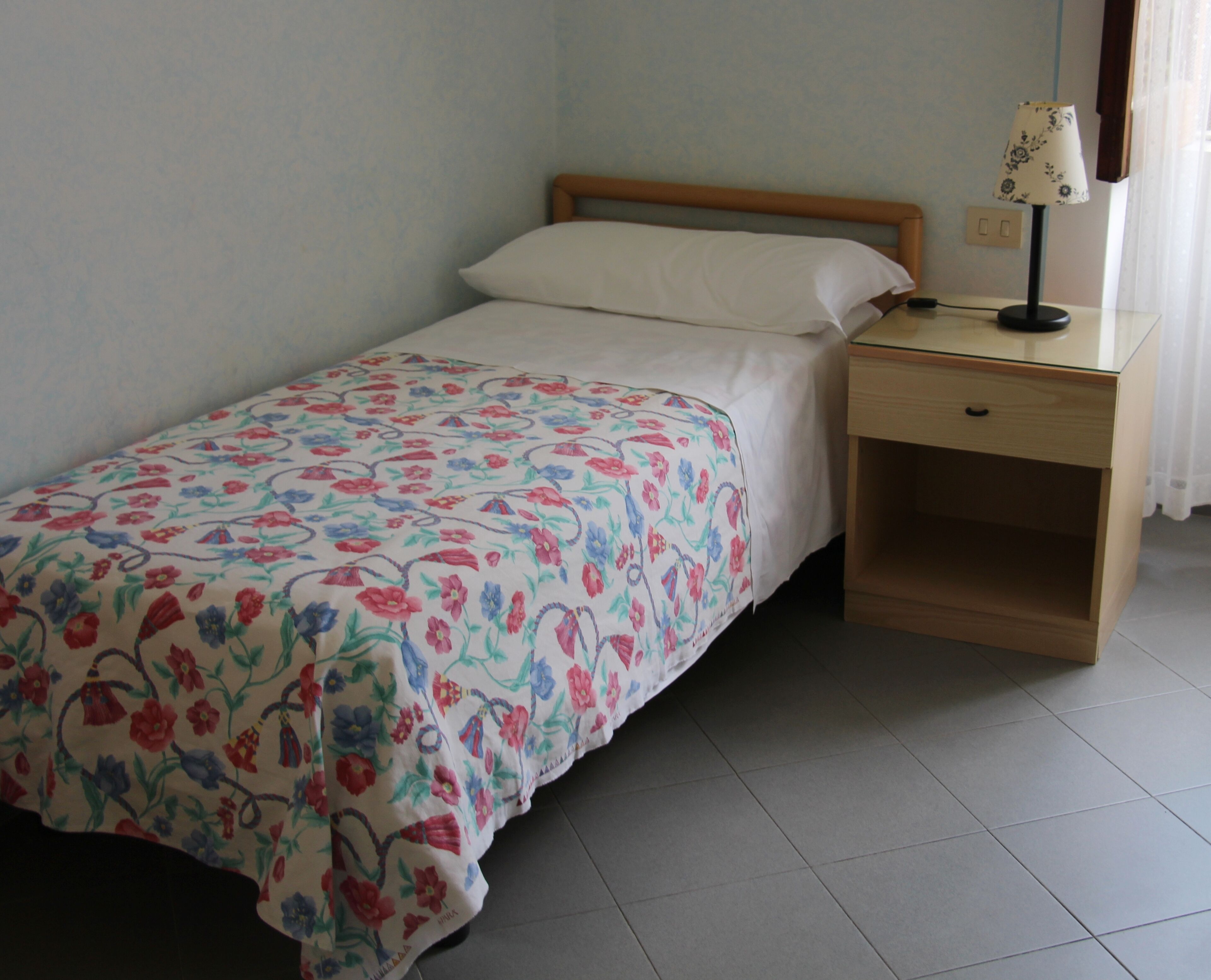 A single bed with a floral bedspread, a wooden headboard, a bedside table with a lamp, and a small drawer.