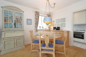 A dining area with a wooden table, blue cushioned chairs, a chandelier, and a cabinet with glass doors. - App. Morgenroth-Munkmarsch (Sylt-Ost)