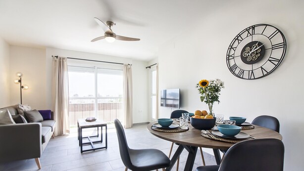 A modern living room with a dining area, a ceiling fan, a large wall clock, and a view through the window. - SW Bailen Beautiful views (Málaga)