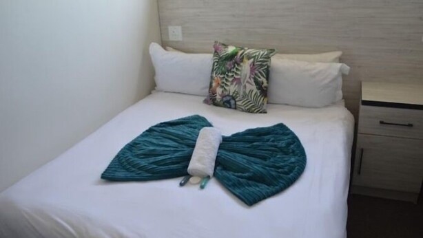 Un lit avec un drap blanc, un coussin décoratif orné d ’un motif de feuilles tropicales et un coussin en forme de nœud de couleur turquoise.