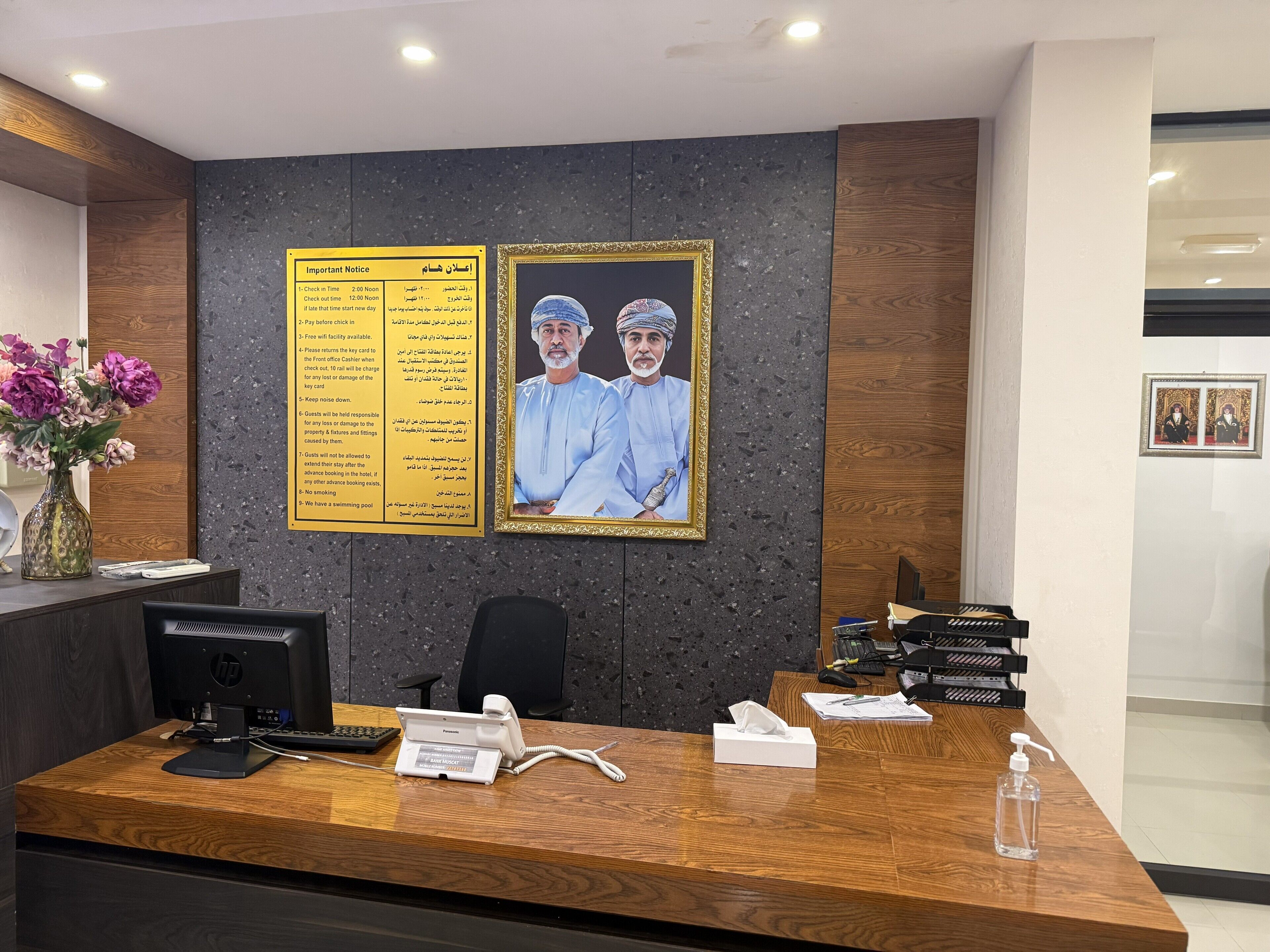 A hotel reception desk with a wooden finish, a Panasonic telephone, a computer, and a vase with flowers.