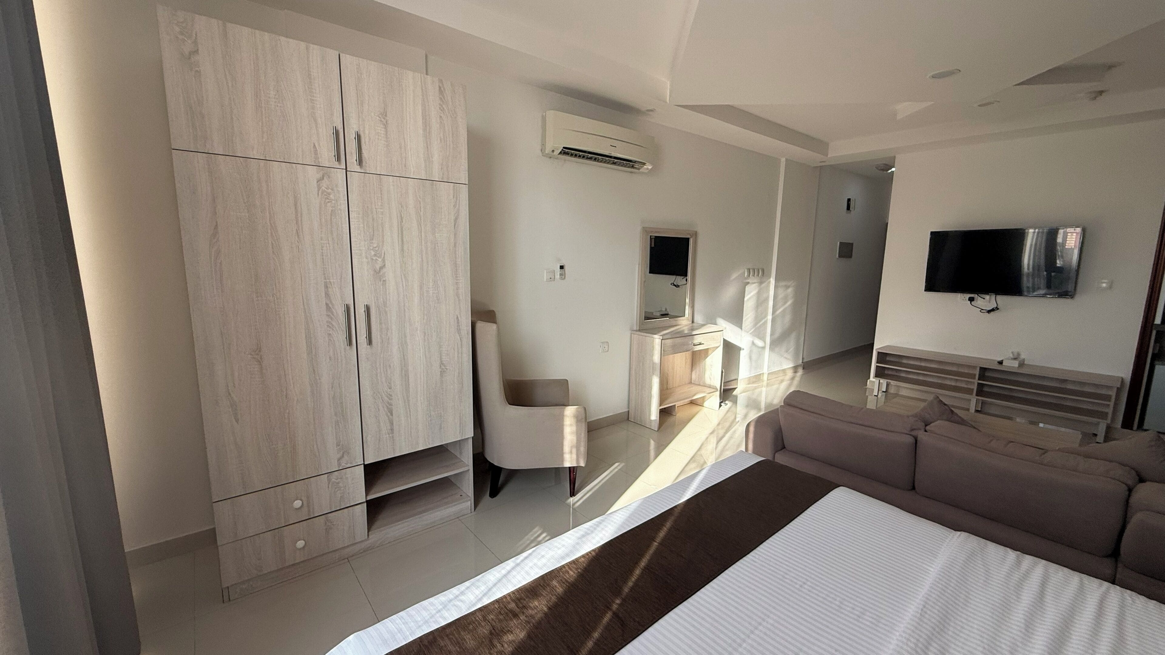 Ein modernes Hotelzimmer mit großem Kleiderschrank, Schreibtisch mit Spiegel, einer Couch und einem wandmontierten Flachbildfernseher.
