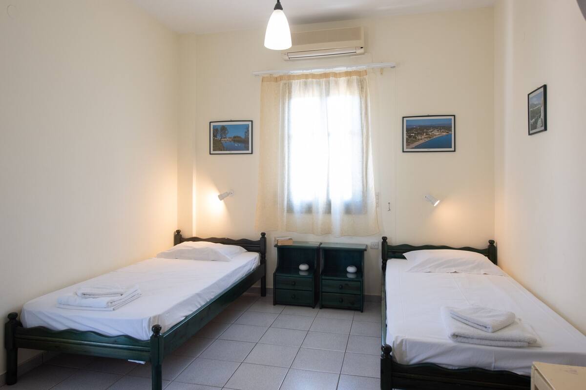 A room with two single beds, a window with curtains, and a wall-mounted air conditioner.