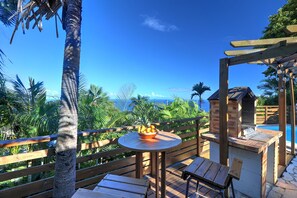 Une terrasse en bois avec une table ronde et des chaises, un petit barbecue en briques, et une vue sur une piscine et des palmiers.