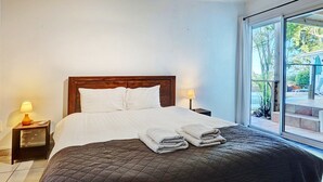Un lit bien fait, avec du linge de lit blanc et une couverture sombre, deux lampes de chevet et une vue sur un balcon agrémenté de plantes et donnant sur une voiture garée à l’extérieur.
