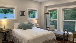 Une chambre à coucher comprenant un lit, des tables de chevet, une lampe, un ventilateur de plafond et de grandes fenêtres donnant sur un paysage verdoyant.