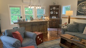 Un salon chaleureux avec un canapé gris, un pouf rouge et une table basse en bois. On y trouve également un coin cuisine avec un tabouret de bar et un meuble en bois.