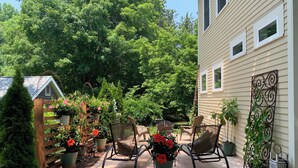 Une terrasse avec des chaises et des plantes en pot, une clôture en bois et une maison dotée d’un garde-corps en métal.