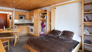 A bedroom with a wooden bed, a wooden desk, a kitchen area with wooden cabinets, and a bookshelf.