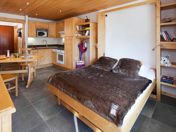 Une chambre avec un lit en bois, un bureau en bois, un coin cuisine avec des placards en bois et une bibliothèque.