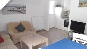 A living room with a sofa, a coffee table, a TV, and a kitchen area with cabinets and appliances.