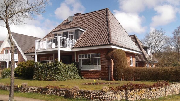 A two-story house with a balcony, brick exterior, and a stone wall in front.