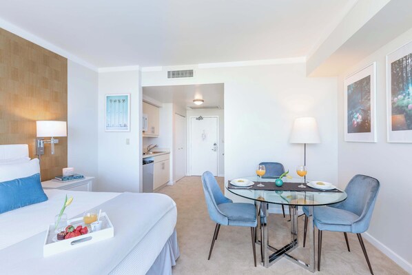 A hotel room with a bed, a dining area with a glass table and chairs, and a kitchenette in the background.