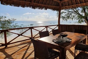 A wooden deck with a table set for dining, overlooking a beach with boats and clear skies.