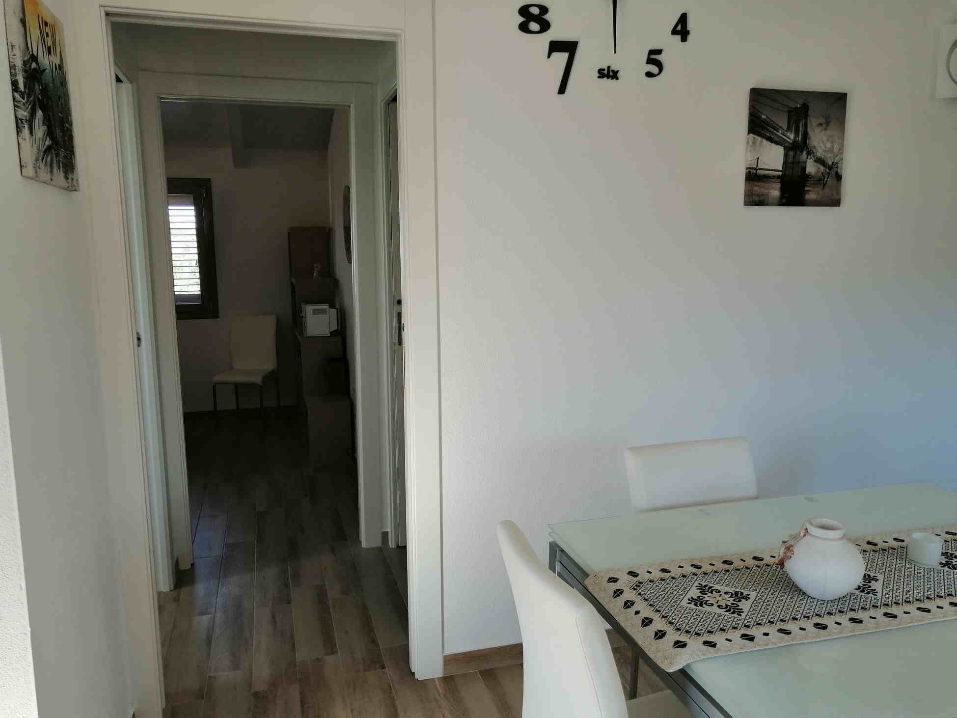 A dining area with a table set for two, a white chair, and a wall clock. A doorway leads to another room.