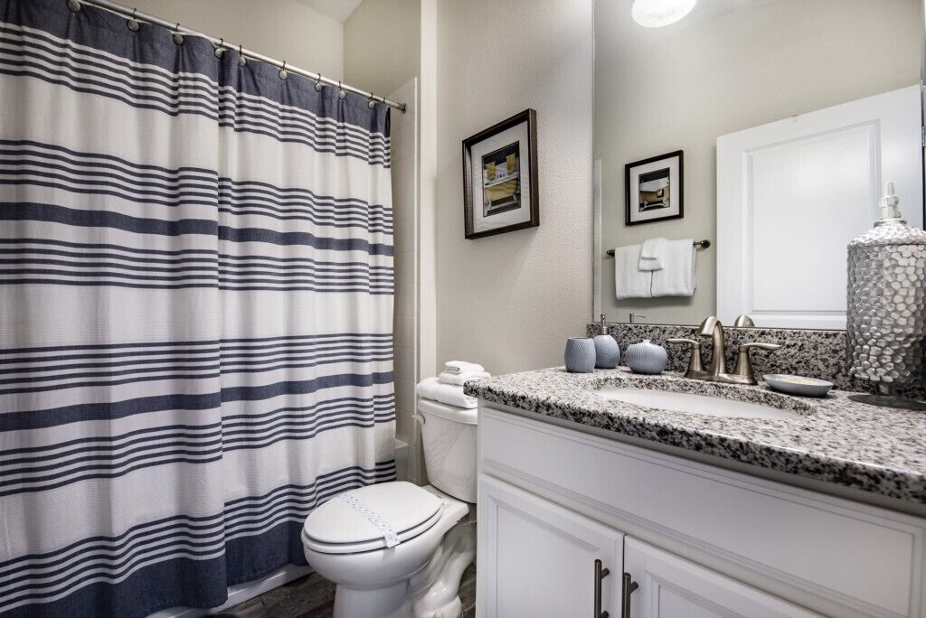 A bathroom with a striped shower curtain, a toilet, a sink with granite countertop, and a mirror.