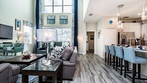 Un'ampia zona living con divano, tavolino e televisione. C'è una sala da pranzo con bancone e sedie. La stanza ha un'ampia finestra e un lampadario.