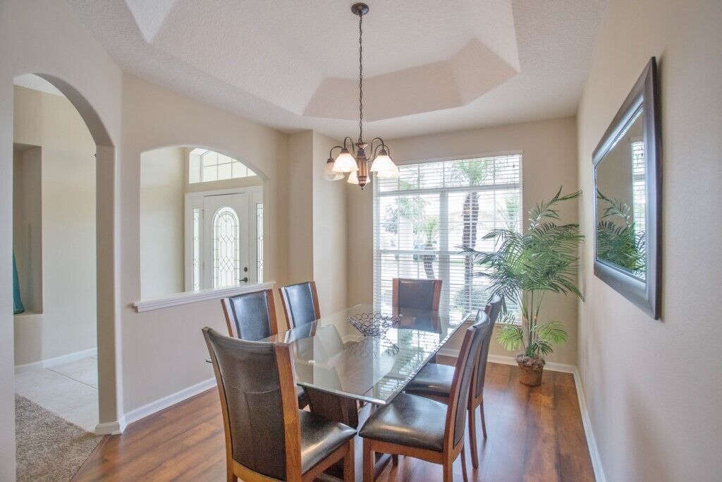 A dining area with a glass table and leather chairs, a chandelier, and a large window with blinds.
