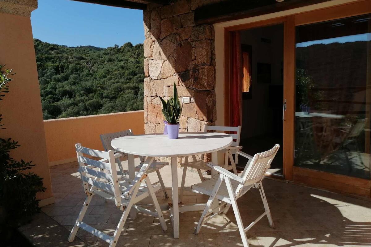 A round table with white chairs and a potted plant on a balcony with stone walls, overlooking a green landscape.
