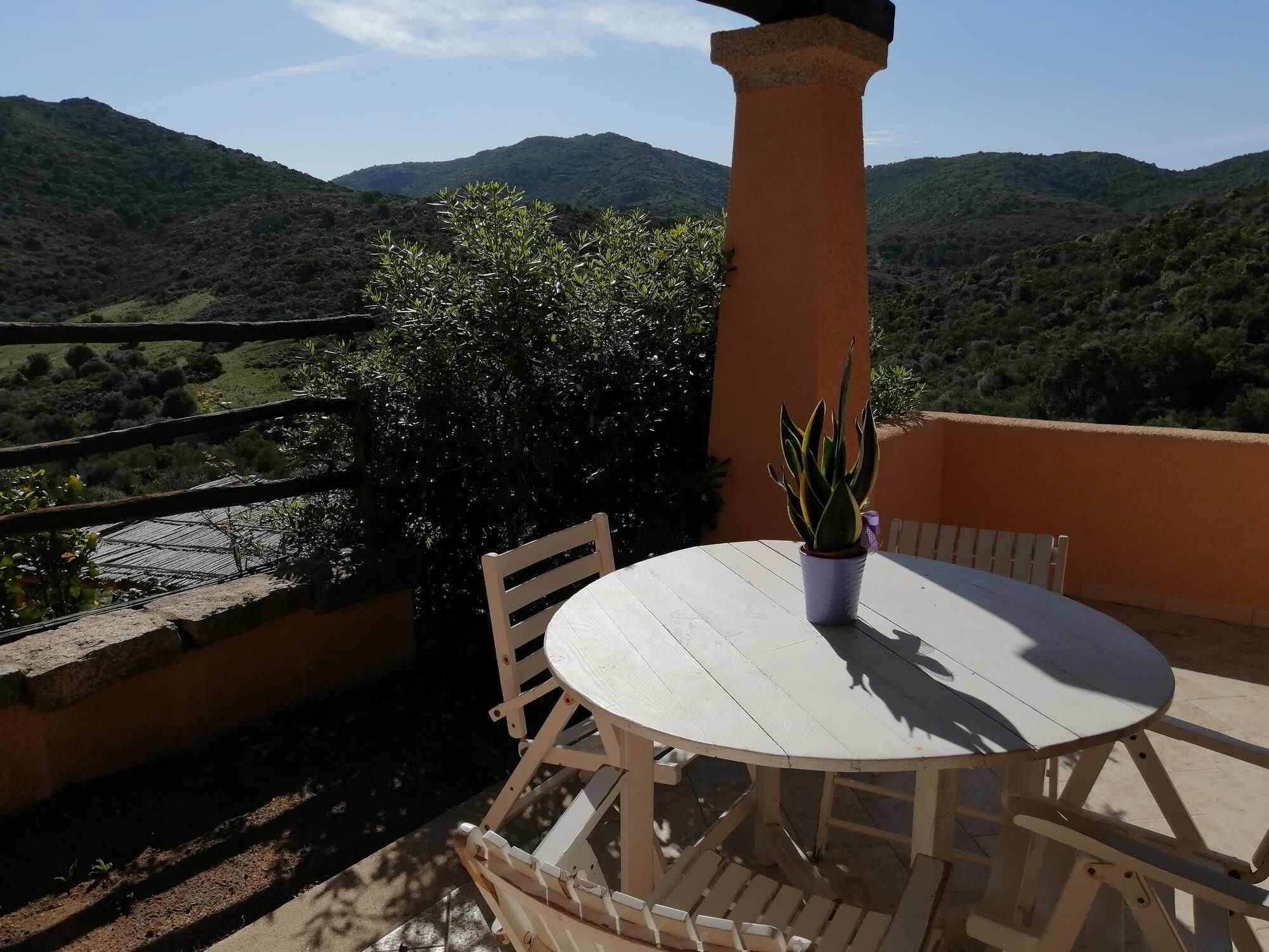 A round wooden table with chairs, a potted plant, and a stone pillar, set against a backdrop of green hills and a clear sky.