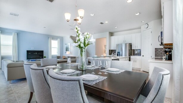 A modern kitchen with white cabinets, a dining area with a dark wood table and grey upholstered chairs, and a living room with a beige sofa and a flat-screen TV. - 1791spp 5 Bedroom Townhouse by RedAwning (Kissimmee)
