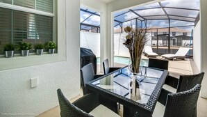 A glass-top dining table with black wicker chairs, a vase with dried plants, and a view of a swimming pool and buildings in the background. - Gorgeous Townhome In Champion's Gate Near Disney! 4 Bedroom Townhouse by RedAwning (Davenport)