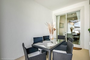 Uma área de refeições com mesa de vidro e cadeiras de vime, um vaso com plantas secas e uma porta de correr de vidro que dá para uma cozinha.