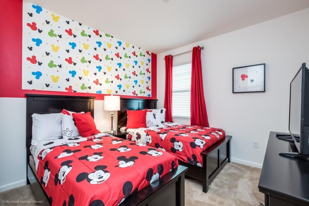 A hotel room with two beds, each with red bedding featuring cartoon characters, a wall with colorful cartoon character decals, a desk with a television, and a window with red curtains.