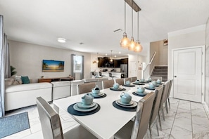 A modern kitchen with dark cabinets, a dining area with a white table set for six, and a staircase leading to an upper level. - 1521MC Amazing Champions Gate 8 Bedroom 5 Bed Villa by RedAwning (Davenport)