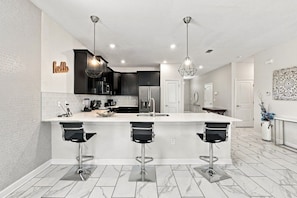 A modern kitchen with a white island, black bar stools, and stainless steel appliances. - 1521MC Amazing Champions Gate 8 Bedroom 5 Bed Villa by RedAwning (Davenport)