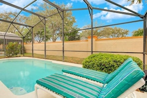 A pool area with a covered section, green-striped lounge chairs, and a view of trees and a building in the background. - Southern Dunes Gated With Private Pool! 4 Bedroom Home by RedAwning (Haines City)