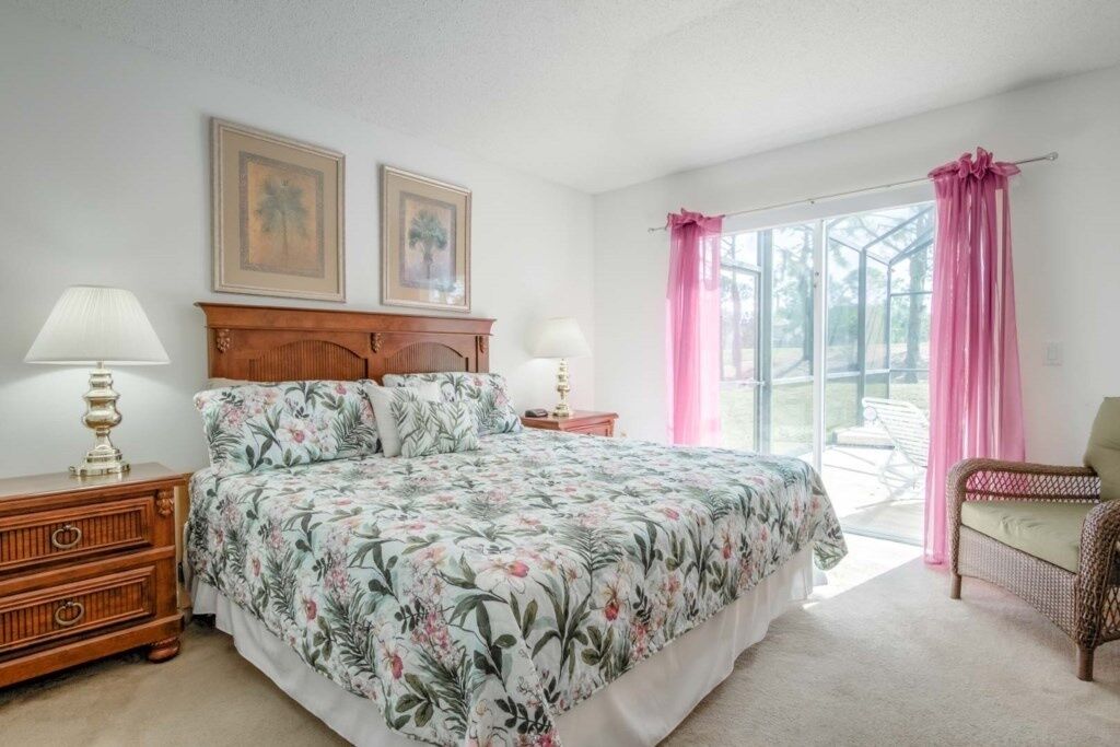 A bedroom with a floral bedspread, two bedside tables with lamps, a wooden dresser, a wicker chair, and a large window with pink curtains.