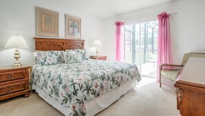 Une chambre à coucher avec une couvre-lit à motifs floraux, une tête de lit en bois, deux lampes de chevet, une commode en bois et une fenêtre avec des rideaux roses.
