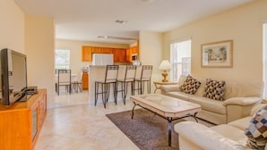 A living room with a sofa, coffee table, and TV stand. There is a kitchen in the background with wooden cabinets and a dining area with chairs.