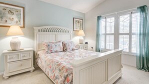 Une chambre à coucher avec un lit blanc, une literie à motifs floraux, deux lampes de chevet, une table de chevet et une fenêtre avec des rideaux.