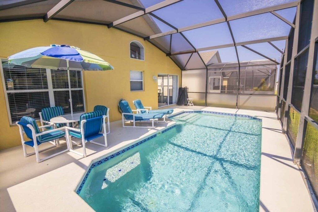 A covered swimming pool with lounge chairs and a table, surrounded by a yellow wall and a glass enclosure.