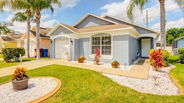 A single-story house with a front garage, a covered patio, and a well-maintained lawn with palm trees and shrubs.