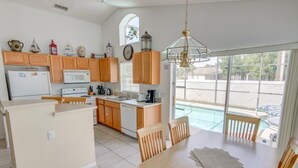 A kitchen with wooden cabinets, a white refrigerator, a microwave, and a coffee maker. There is a dining table with chairs and a view of a swimming pool through large windows.