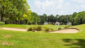 Ein Golfplatz mit Sandbunkern, einem gepflegten Rasen und einer Reihe von Häusern im Hintergrund.