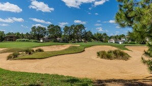 A golf course with sand bunkers, a clear sky, and residential houses in the background.