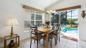 A dining area with a wooden table, six chairs, a vase with flowers, a lamp, and a view of a swimming pool through large windows.