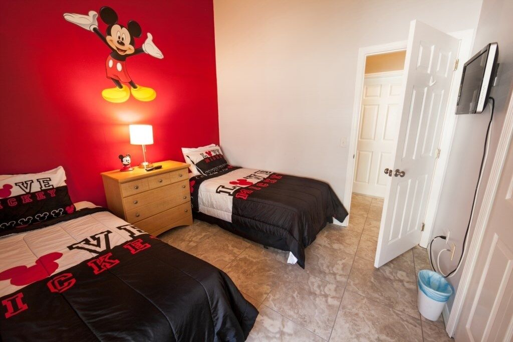Ein Schlafzimmer mit zwei Betten, einem Schminktisch, einer Wanddekoration mit Mickey Mouse, einem Fernseher und einer Tür zu einem anderen Raum.