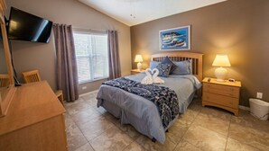A bedroom with a bed, two bedside tables, a TV mounted on the wall, a window with curtains, and a ceiling fan.