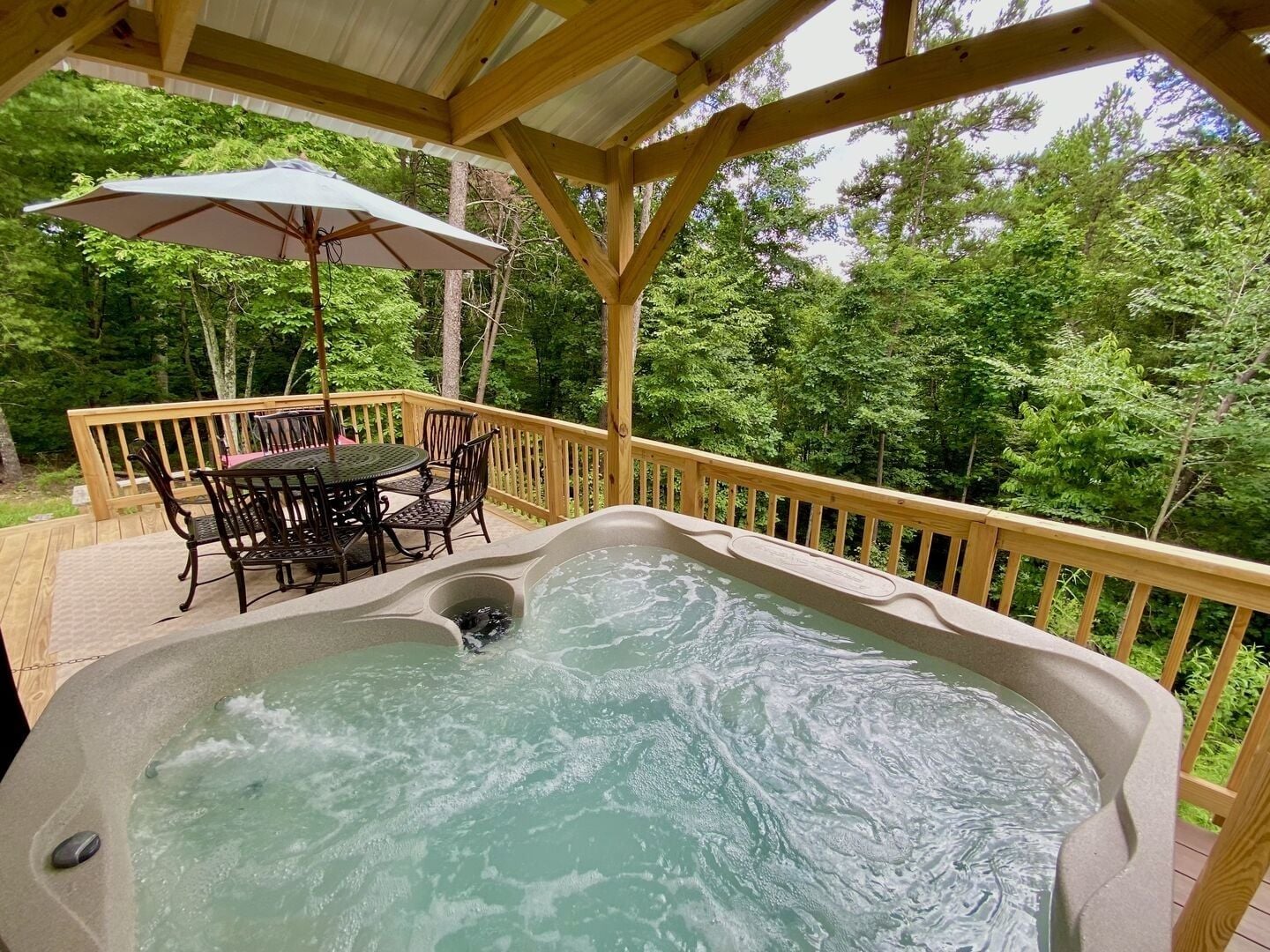 A hot tub with a wooden deck, surrounded by trees, and a table with chairs under a canopy.