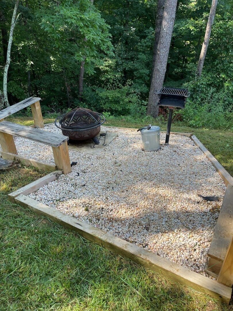 A gravel area with a metal fire pit, a wooden bench, and a bird feeder.