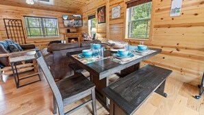 A cozy cabin interior with a wooden dining table set for four, a fireplace, and a large window overlooking greenery. - Foxes' Den Cabin with Private Hot Tub and Arcade Game by RedAwning (Pigeon Forge)